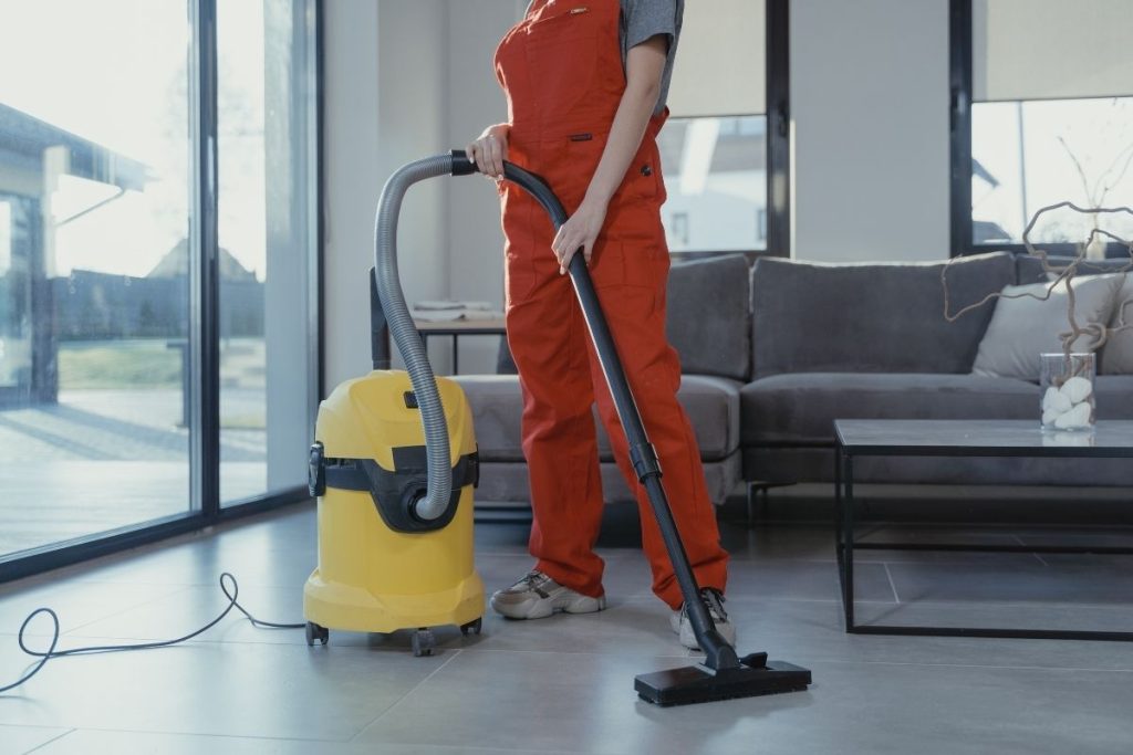 A woman wearing an orange overall stands next to a vacuum cleaner, preparing to start her cleaning task.